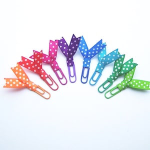 May include: A collection of colorful paper clips, each adorned with a polka-dotted ribbon bow. The bows are in a rainbow of colors, including orange, red, pink, purple, blue, teal, and green. The paper clips are in matching colors.