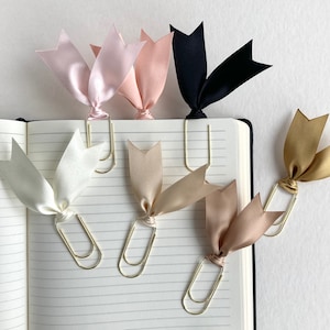 May include: Five gold paper clips with different coloured satin ribbon bows. The bows are in pink, white, beige, gold, and black.