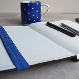 May include: An open notebook with a blue elastic band closure, two pens, and a cell phone on a wooden surface.