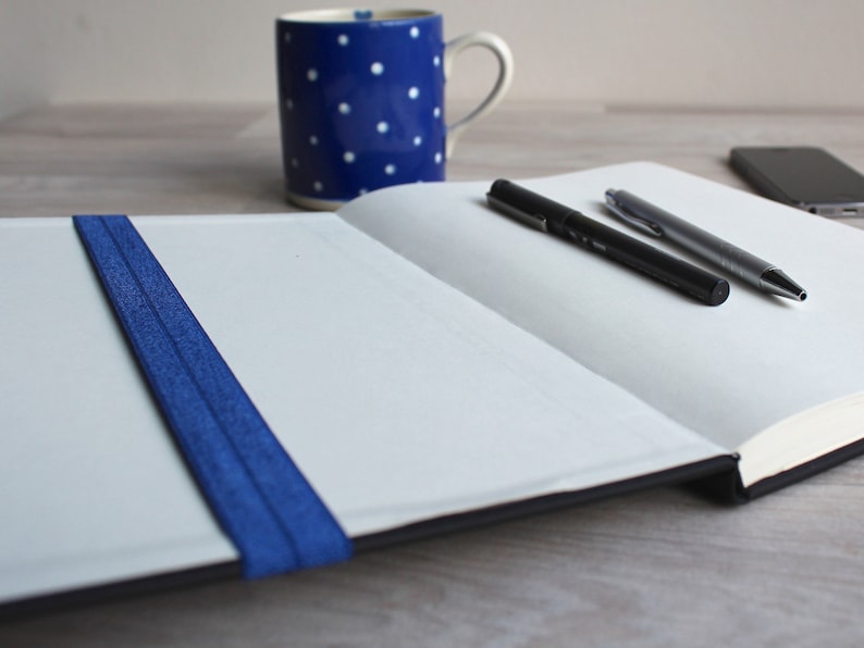 May include: An open notebook with a blue elastic band closure, two pens, and a cell phone on a wooden surface.