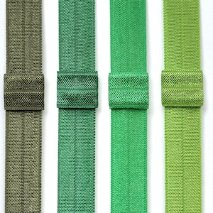 May include: A close-up of four elastic bands in shades of green and olive. Each band has a matching fabric-covered loop. The colours range from a muted olive green to a bright, vibrant green. The bands are arranged vertically against a white background.
