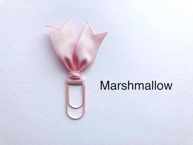 Może przedstawiać: R&oacute;żowa wstążka zawiązana wok&oacute;ł r&oacute;żowego złotego spinacza. Wstążka jest zawiązana w kokardkę. Tekst "Marshmallow" jest napisany pod spinaczem.