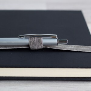 May include: A black leather bound journal with a silver pen secured by a gray ribbon.