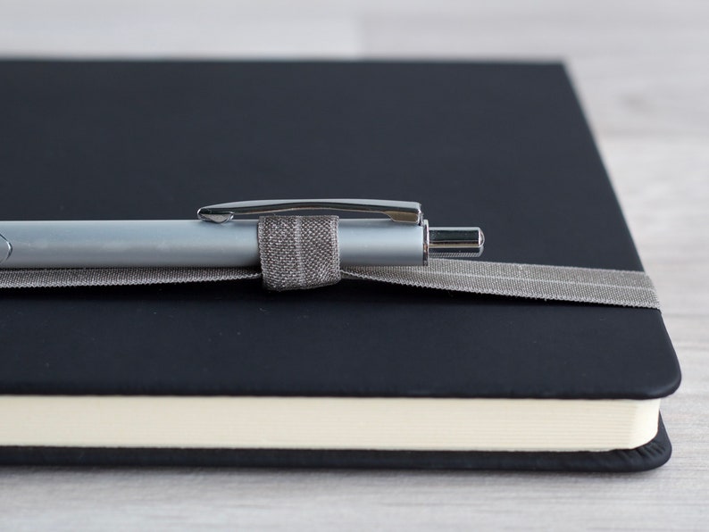 May include: A black leather bound journal with a silver pen secured by a gray ribbon.