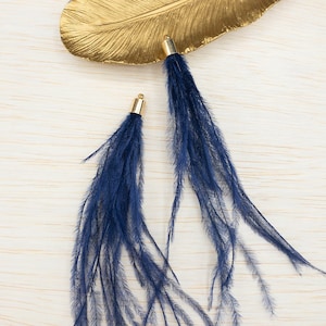 May include: Two blue feather trim tassels with gold metal caps. The tassels are on a light wood surface.