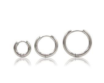 May include: Three silver-toned hoop earrings of varying sizes arranged in a horizontal row. Each earring features a smooth, polished metallic finish and a hinged closure mechanism, creating a simple circular design with a reflective surface.