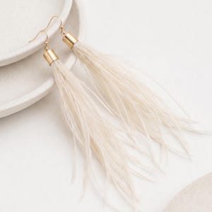 Puede incluir: Un par de pendientes largos de plumas color crema con detalles met&aacute;licos dorados. Los pendientes se muestran sobre una superficie blanca. El texto en la parte inferior dice: Subproductos de origen &eacute;tico de la industria de la confecci&oacute;n.