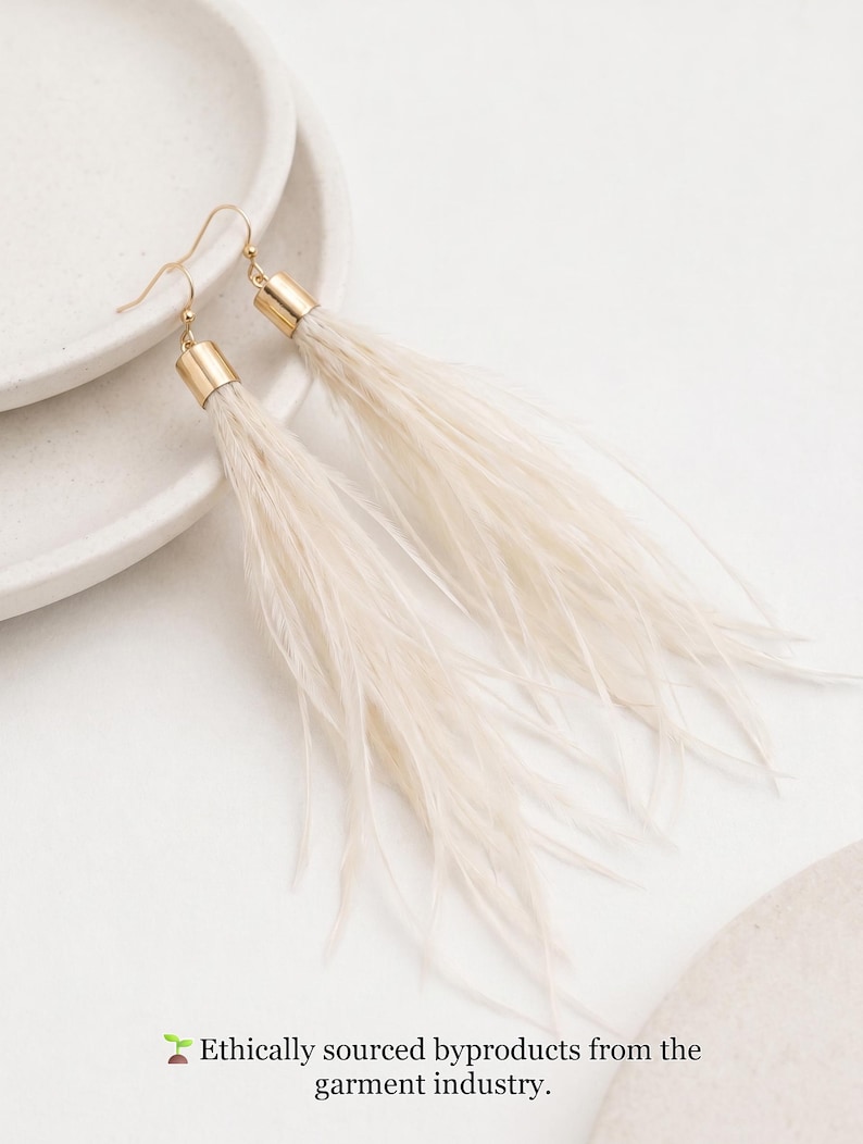 Puede incluir: Un par de pendientes largos de plumas color crema con detalles met&aacute;licos dorados. Los pendientes se muestran sobre una superficie blanca. El texto en la parte inferior dice: Subproductos de origen &eacute;tico de la industria de la confecci&oacute;n.
