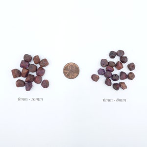 May include: Two groups of irregular, dark reddish-purple gemstone beads arranged on a white surface. The left group is labeled 8mm - 10mm, and the right group is labeled 6mm - 8mm. A copper-colored United States penny is placed between the two groups.