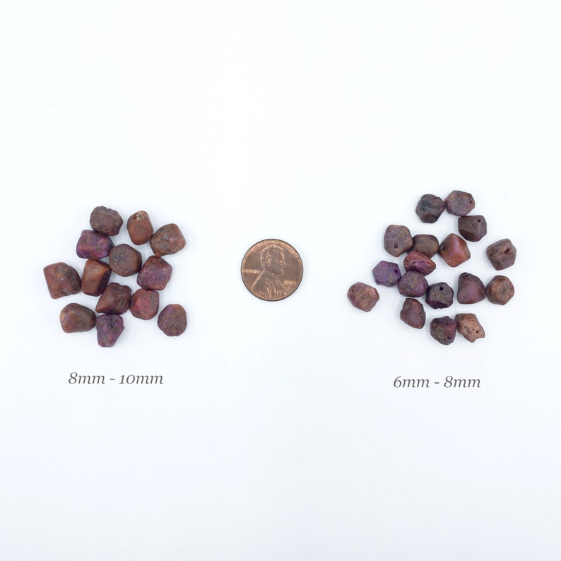 May include: Two groups of irregular, dark reddish-purple gemstone beads arranged on a white surface. The left group is labeled 8mm - 10mm, and the right group is labeled 6mm - 8mm. A copper-colored United States penny is placed between the two groups.