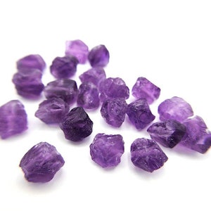 May include: A collection of small, raw amethyst gemstones with irregular, jagged shapes and translucent purple coloring. Each stone features natural facets and varying shades of deep violet, scattered across a plain white surface.