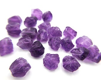 May include: A collection of small, raw amethyst gemstones with irregular, jagged shapes and translucent purple coloring. Each stone features natural facets and varying shades of deep violet, scattered across a plain white surface.