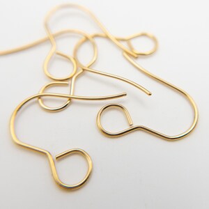 Peut inclure: Une collection de crochets de boucles d'oreilles en m&eacute;tal dor&eacute;, chacun pr&eacute;sentant un design en fil courb&eacute; avec une petite boucle circulaire &agrave; la base pour attacher des &eacute;l&eacute;ments de bijoux. Ces appr&ecirc;ts sont dispos&eacute;s en tas.