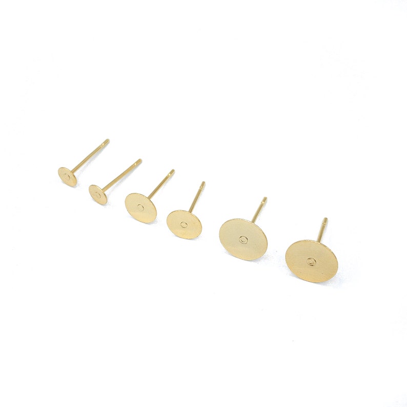 May include: Six gold-toned metal earring studs arranged in a row, increasing in size from left to right. Each stud features a flat, circular pad attached to a thin post, designed for jewelry making and attaching decorative elements to the front.