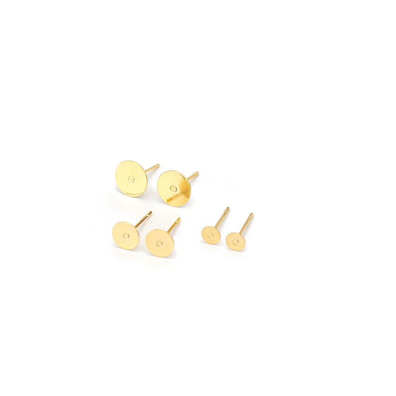 May include: A collection of six gold-toned stud earring components featuring flat circular pads for attaching decorations. The set includes two large studs, two medium studs, and two small studs, each with a thin post extending from the back.