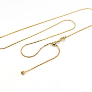 May include: A gold-toned snake chain necklace featuring a sliding adjustable bead mechanism. The chain has a smooth, flexible texture and terminates in a small, polished gold-toned sphere at the end of the adjustable section, secured by a spring ring clasp.