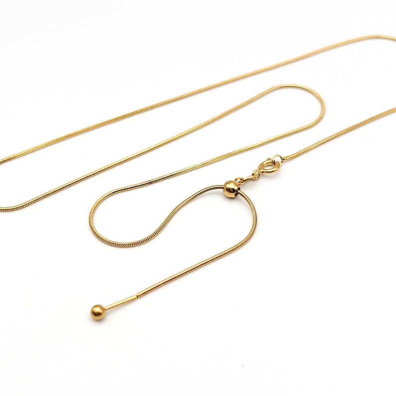May include: A gold-toned snake chain necklace featuring a sliding adjustable bead mechanism. The chain has a smooth, flexible texture and terminates in a small, polished gold-toned sphere at the end of the adjustable section, secured by a spring ring clasp.
