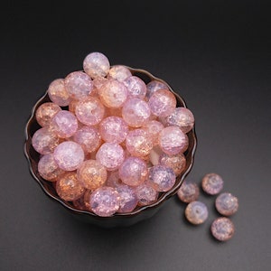 May include: A bowl filled with pink and white iridescent glass beads. The beads are round and have a textured surface. There are also a few beads scattered on the table.