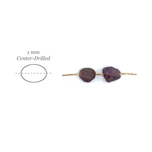 May include: Two raw, deep red ruby beads threaded on a thin gold wire. A diagram to the left shows an oval shape with a dashed line through the center, labeled 1 mm Center-Drilled, indicating the hole placement for these natural gemstone beads.