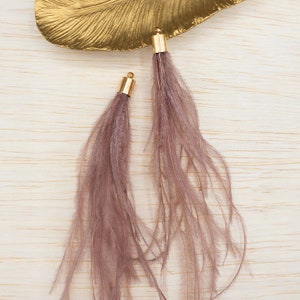 May include: Two feather boas with gold metal tips. The boas are a dusty rose color and are laying on a light wood surface.