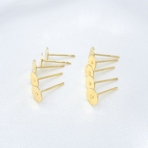 May include: A collection of gold-toned metal earring studs, each featuring a flat, circular pad at the top for attaching decorative elements and a straight post extending from the center, arranged in two rows on a white surface.
