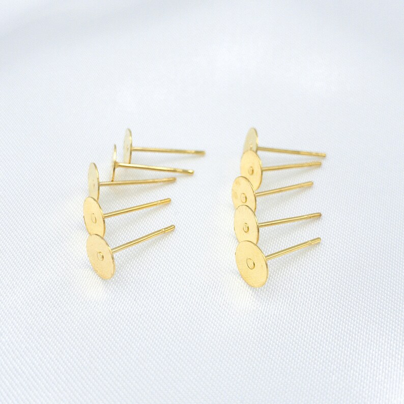 May include: A collection of gold-toned metal earring studs, each featuring a flat, circular pad at the top for attaching decorative elements and a straight post extending from the center, arranged in two rows on a white surface.