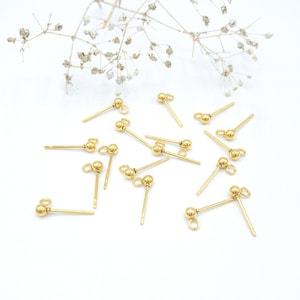 May include: A group of 14 gold-toned earring posts with a small ball at the top and a loop at the bottom. The earring posts are scattered on a white surface.