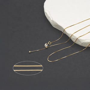 May include: A gold-toned box chain necklace featuring a sliding adjustable mechanism with a small round bead and a thin metal tip. The chain consists of square-shaped links, and a small inset shows a magnified view of the chain's square link pattern.