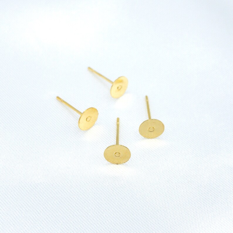 May include: Four gold-toned metal earring posts with flat circular pads. Each component features a thin, straight pin attached to the center of a small, round, textured disc, designed for attaching decorative elements to create stud earrings.