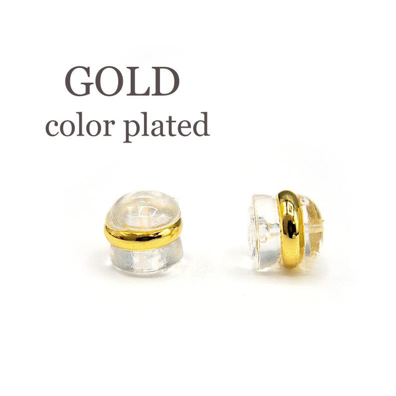 Puede incluir: Dos respaldos de pendientes chapados en oro, cada uno con un respaldo de pl&aacute;stico transparente y un anillo dorado alrededor del borde. El texto "GOLD color plated" es visible sobre los respaldos de los pendientes.