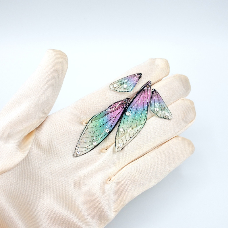 May include: A set of three iridescent dragonfly wing earrings with black veins and clear rhinestones. The earrings are displayed on a white fabric background.