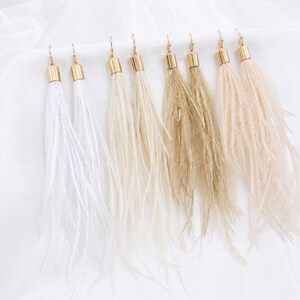 Puede incluir: Una colecci&oacute;n de pendientes de plumas en varios colores neutros, incluyendo blanco, beige y marr&oacute;n. Cada pendiente presenta una tapa y un gancho de metal dorado. Las plumas caen en cascada, creando un aspecto fluido y elegante. Los pendientes se muestran sobre un fondo blanco.