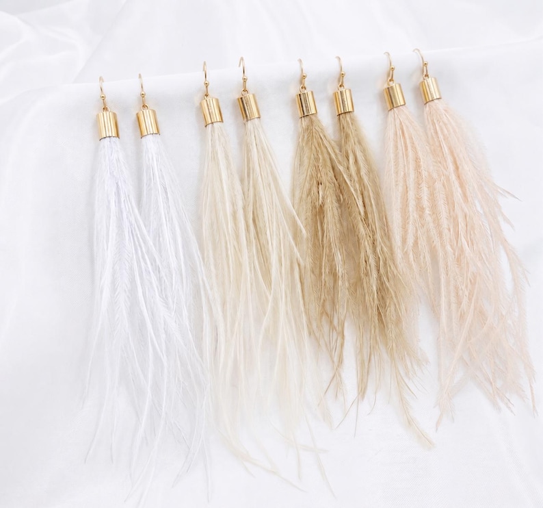 Puede incluir: Una colecci&oacute;n de pendientes de plumas en varios colores neutros, incluyendo blanco, beige y marr&oacute;n. Cada pendiente presenta una tapa y un gancho de metal dorado. Las plumas caen en cascada, creando un aspecto fluido y elegante. Los pendientes se muestran sobre un fondo blanco.
