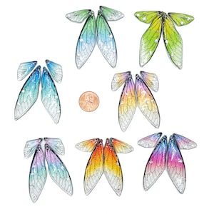 May include: Set of six pairs of iridescent dragonfly wings, each pair featuring a different color gradient. The wings are made of a translucent material with a delicate vein pattern and glitter accents. The wings are arranged in a grid pattern on a white background.