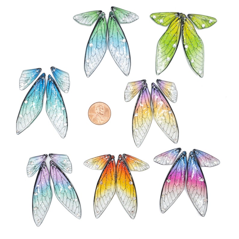 May include: Set of six pairs of iridescent dragonfly wings, each pair featuring a different color gradient. The wings are made of a translucent material with a delicate vein pattern and glitter accents. The wings are arranged in a grid pattern on a white background.
