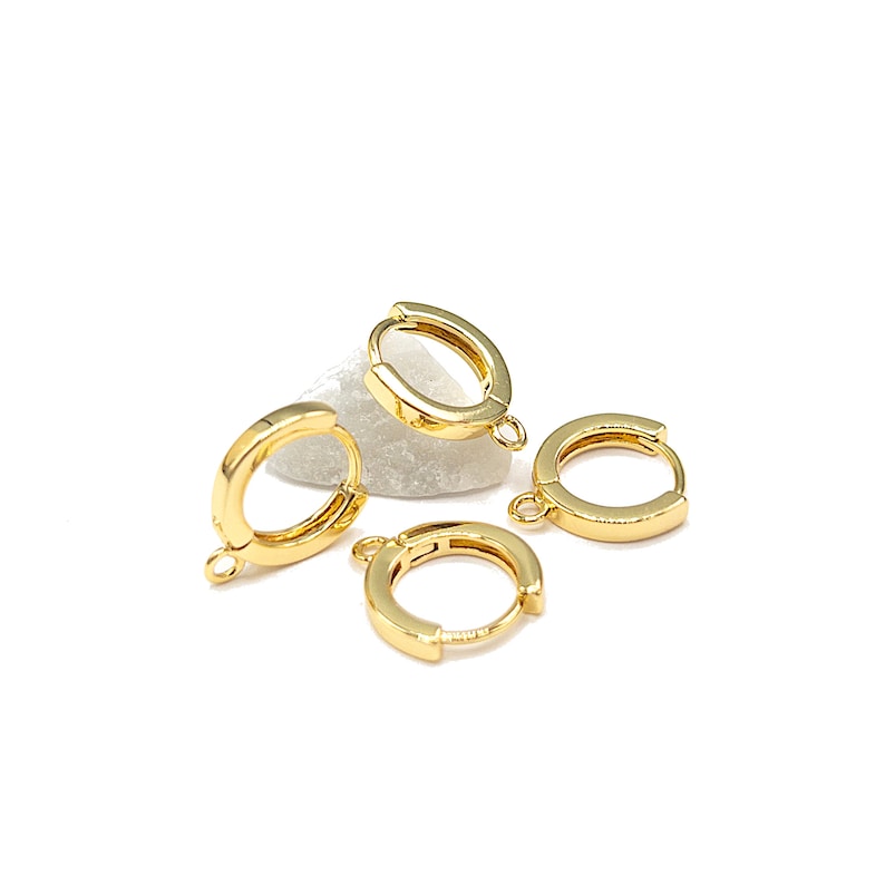 May include: Four small, polished gold-tone hoop earrings, each featuring a hinged closure and a tiny circular loop at the bottom for attaching charms. The hoops are arranged in a cluster, showcasing their smooth, reflective metallic finish.