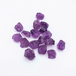 Peut inclure: Une collection de petites pierres pr&eacute;cieuses d'am&eacute;thyste brutes aux formes irr&eacute;guli&egrave;res et angulaires, pr&eacute;sentant des nuances vari&eacute;es de violet profond. Chaque pierre poss&egrave;de une texture cristalline translucide avec des facettes naturelles.