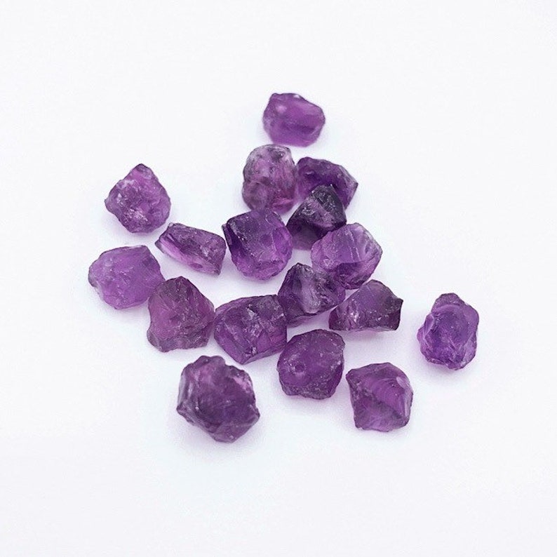 Peut inclure: Une collection de petites pierres pr&eacute;cieuses d'am&eacute;thyste brutes aux formes irr&eacute;guli&egrave;res et angulaires, pr&eacute;sentant des nuances vari&eacute;es de violet profond. Chaque pierre poss&egrave;de une texture cristalline translucide avec des facettes naturelles.