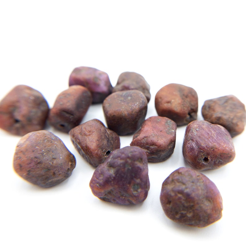 May include: A collection of twelve small, irregularly shaped beads with a rough, textured surface. Each bead displays a deep, earthy reddish-brown color with subtle purple undertones and a single drilled hole, showcasing a natural, unpolished appearance.
