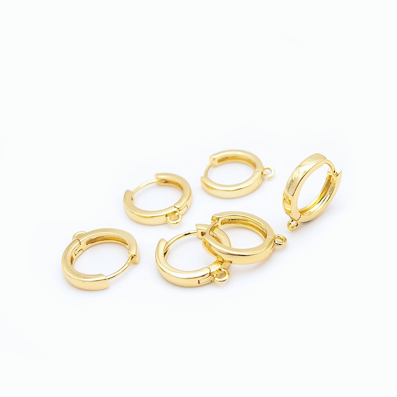 Op de afbeelding: Een verzameling van vijf kleine, gepolijste goudkleurige oorringen, elk voorzien van een scharniersluiting en een klein ge&iuml;ntegreerd oogje aan de onderkant voor het bevestigen van bedels of andere sieraden.
