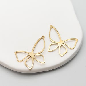 May include: Two gold-colored butterfly-shaped charms. Each charm features a delicate, openwork design with curved wings and a small loop at the top for attaching to jewelry. The charms are displayed on a white surface.