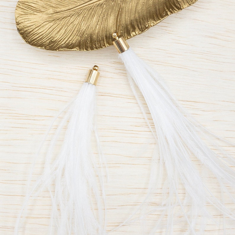 Puede incluir: Dos borlas de plumas blancas con tapas de metal dorado. Las borlas se exhiben sobre una fuente dorada con forma de hoja, sobre un fondo claro de vetas de madera. Las plumas son largas y tenues.