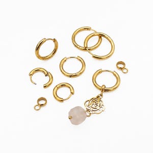 May include: A collection of gold-toned hoop earrings in various sizes, featuring a hinged closure mechanism. One hoop earring includes a dangling charm with a cutout floral design and a single, pale pink, irregular-shaped stone bead attached.