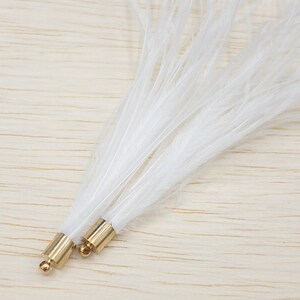 Puede incluir: Primer plano de dos plumas blancas con bases de metal dorado. Las plumas son largas, suaves y delicadas, abri&eacute;ndose en abanico desde la base. Las bases doradas tienen una peque&ntilde;a perla redonda dorada en el extremo. El fondo es de vetas de madera clara.