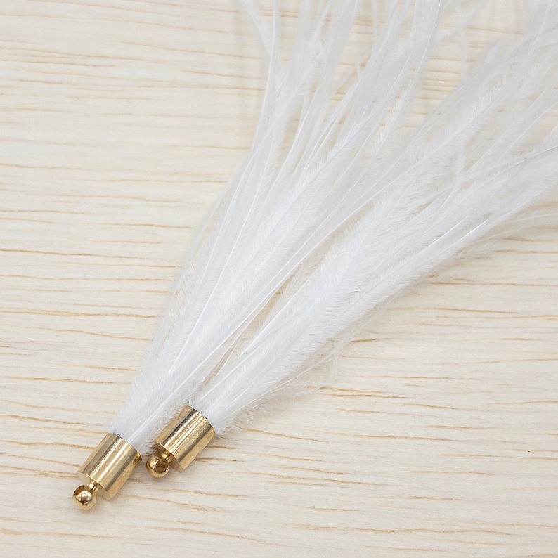 Puede incluir: Primer plano de dos plumas blancas con bases de metal dorado. Las plumas son largas, suaves y delicadas, abri&eacute;ndose en abanico desde la base. Las bases doradas tienen una peque&ntilde;a perla redonda dorada en el extremo. El fondo es de vetas de madera clara.
