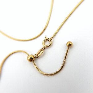May include: A gold-toned snake chain necklace featuring a sliding adjustable mechanism with a small round bead, a spring ring clasp, and a straight metal bar end piece. The chain has a smooth, flexible texture and a polished metallic finish.