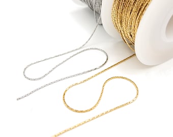 May include: Two spools of metallic chain, one silver and one gold, are shown with lengths of each chain extending outward. The chains feature a textured, repeating link design, and the spools are white plastic with a central core for dispensing.