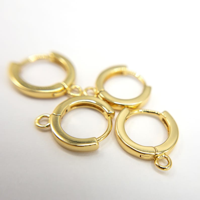 May include: Four gold-toned hoop earrings arranged in a cluster. Each hoop features a smooth, polished finish and a hinged closure mechanism. Two of the hoops include a small, circular loop attached to the bottom edge for hanging additional charms.
