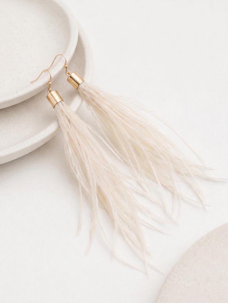 Puede incluir: Un par de pendientes largos de plumas color crema con detalles met&aacute;licos dorados. Los pendientes se exhiben sobre una superficie blanca, mostrando su dise&ntilde;o delicado y fluido. Los pendientes tienen un cierre de gancho.