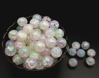 May include: A collection of round, crackled glass beads with an iridescent finish. The beads display a translucent quality with shifting hues of pale blue, soft pink, and white. Many beads feature a central hole, and they are arranged in a bowl.